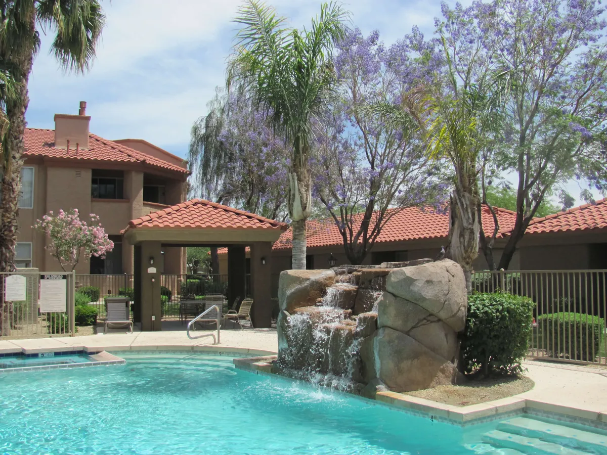 The image showcases a sparkling swimming pool surrounded by lush greenery and vibrant purple flowering trees, with elegant residential buildings as a backdrop. The tranquil setting invites relaxation, sunbathing, and socializing, offering a refreshing oasis in the heart of North Phoenix.