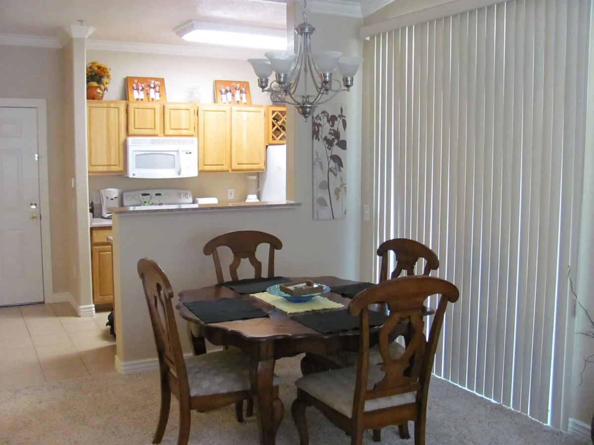 A welcoming dining area anchored by a dining table and chandelier, with kitchen cabinets and sliding blinds visible in the background. The setting suggests a seamless flow between cooking and dining, suitable for family meals or casual gatherings.