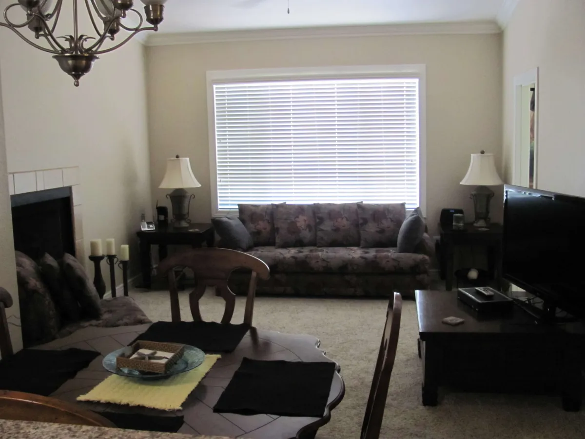 The image displays a cozy living area featuring a floral-patterned sofa, a dining table, elegant chandelier, and a television as a focal point. The space exudes a comfortable and homey ambiance, with multiple seating options inviting relaxation and social gatherings. The TV is prominently placed, making it ideal for movie nights or catching up on shows.
