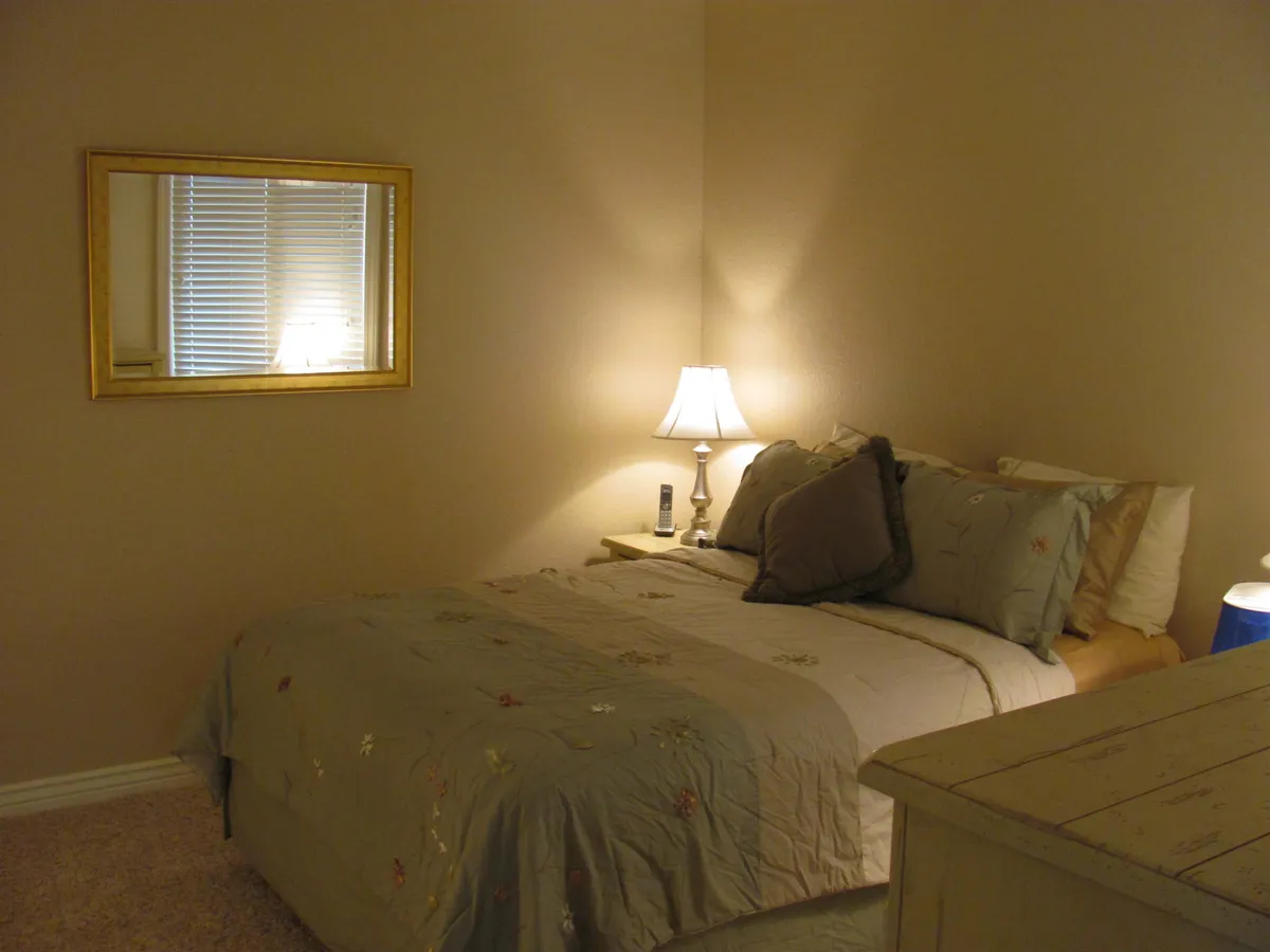 This image features a tranquil bedroom scene with a comfortable bed adorned with decorative pillows, a softly glowing lamp on the nightstand, and a mirror that adds depth to the space. The serene color palette and plush bedding make the room feel inviting and restful, ideal for unwinding after a long day.